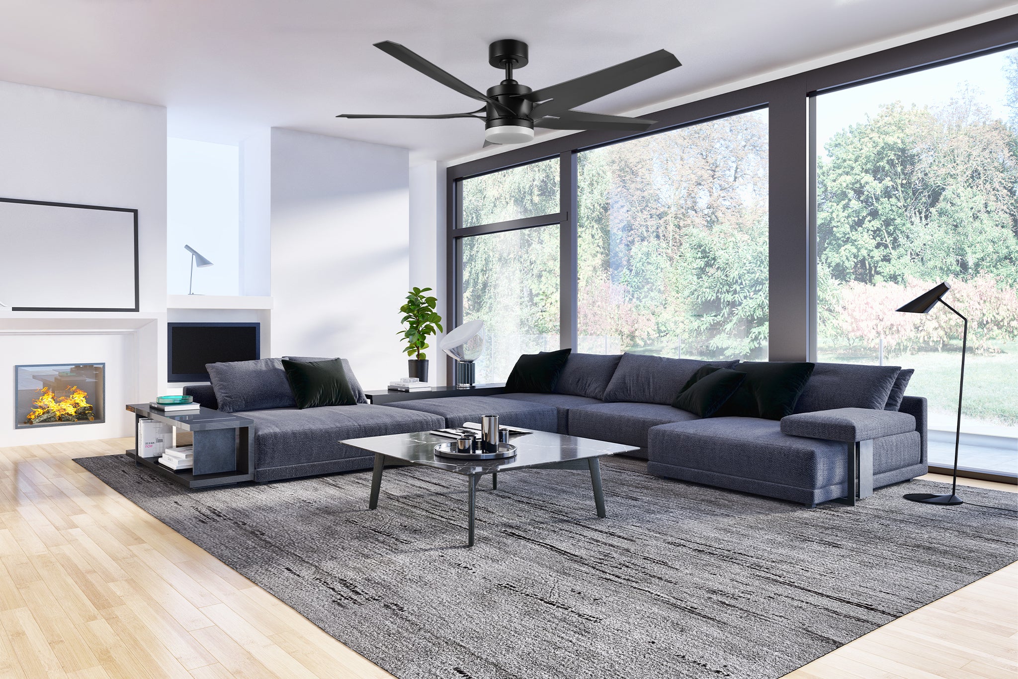 A modern living room featuring a 52-inch Araceli LED lighting ceiling fan with a remote control. The room has large floor-to-ceiling windows showcasing a view of greenery outside. The interior is decorated with a minimalist style, including two dark gray sectionals with black throw pillows, a gray area rug, and light wooden flooring. There is a fireplace on the left wall, a coffee table with decorative items in the center, and a potted plant adding a touch of greenery to the space.