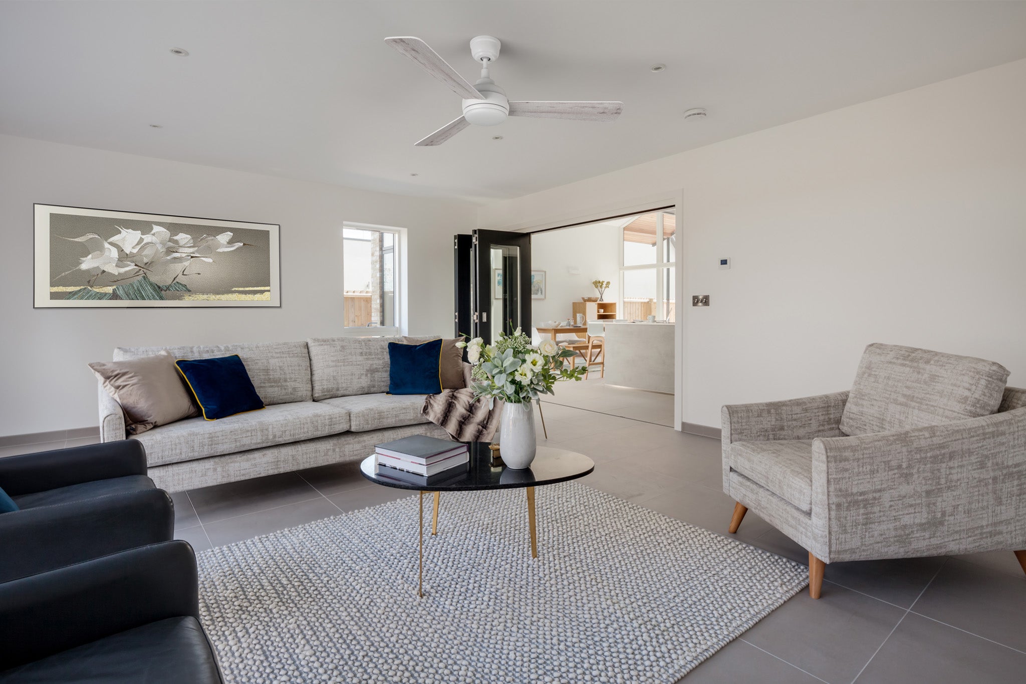 52 inch Audra white ceiling fan with light in a modern living room featuring a light grey sofa with blue and brown pillows, two dark grey armchairs, a patterned rug, and a glass-top coffee table with flowers. A large painting decorates the wall above the sofa.