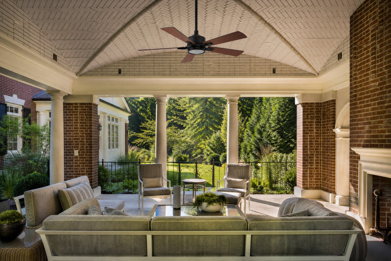 Matte black ceiling fan with light fixture and five medium-toned wood blades installed on a vaulted herringbone ceiling. The outdoor ceiling fan anchors a luxury covered patio space complete with comfortable outdoor furniture, brick pillars, and backyard views.