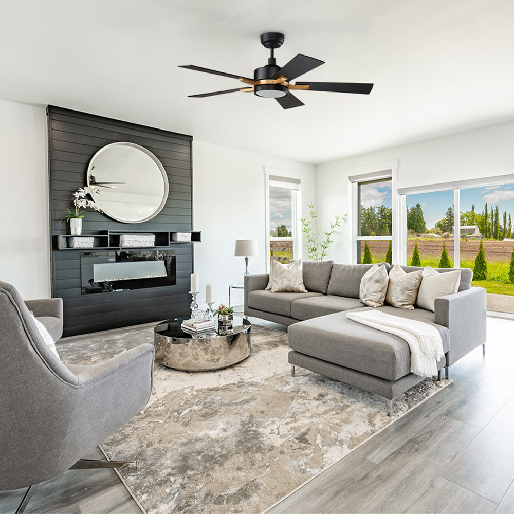A modern, bright living room featuring 52 inch Apex black and gold smart ceiling fan. The room is decorated with a large, gray sectional sofa, a matching armchair, a round silver coffee table, and a rug. A black fireplace and a large round mirror are on one wall, while large windows offer a view of an open field.