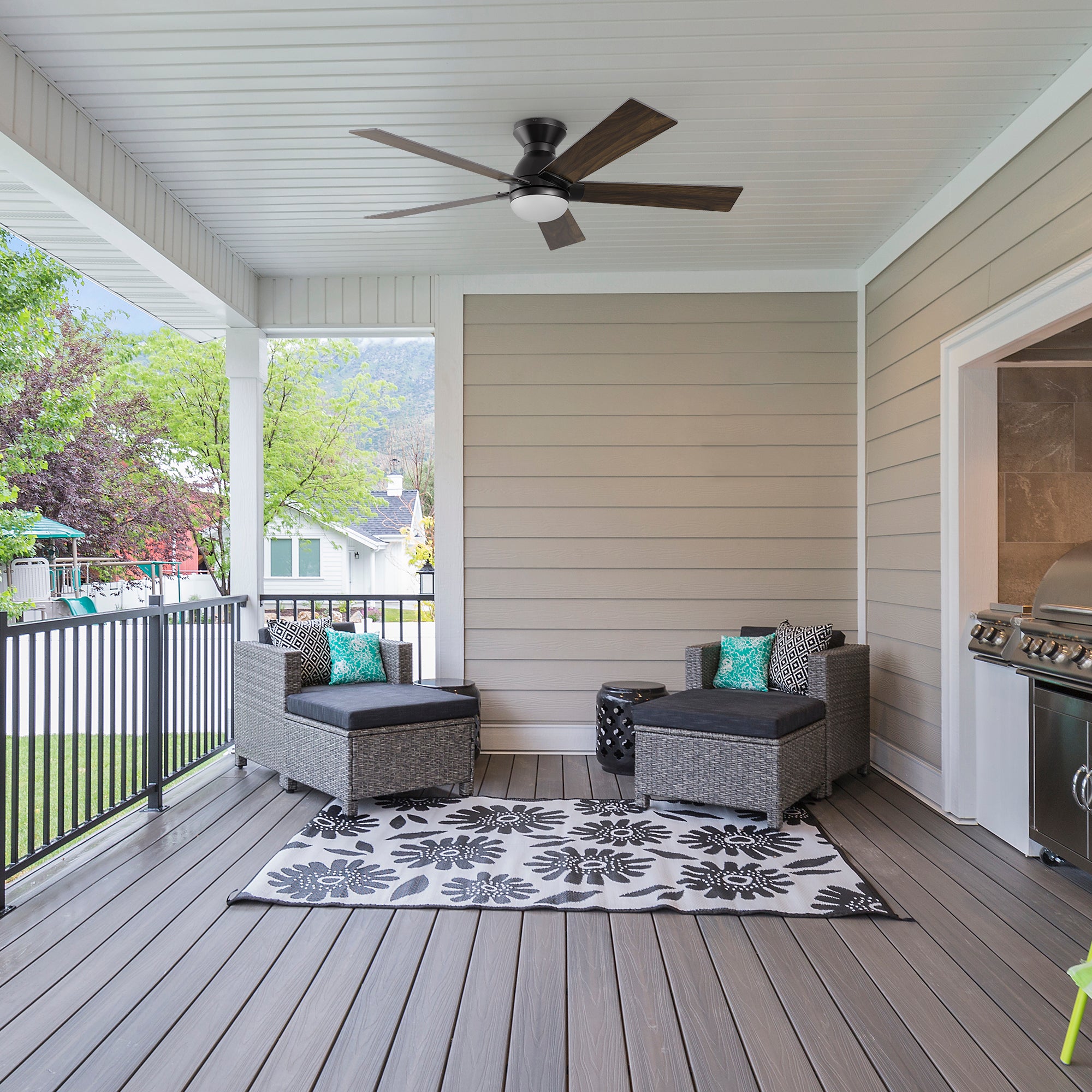Covered wood patio featuring the Aspen 52-inch outdoor low-profile smart ceiling fan with light, mounted on a white paneled ceiling. The space is furnished with gray wicker lounge seating, colorful accent pillows, a black and white floral rug, and a stainless steel grill, offering a cozy and stylish outdoor retreat.