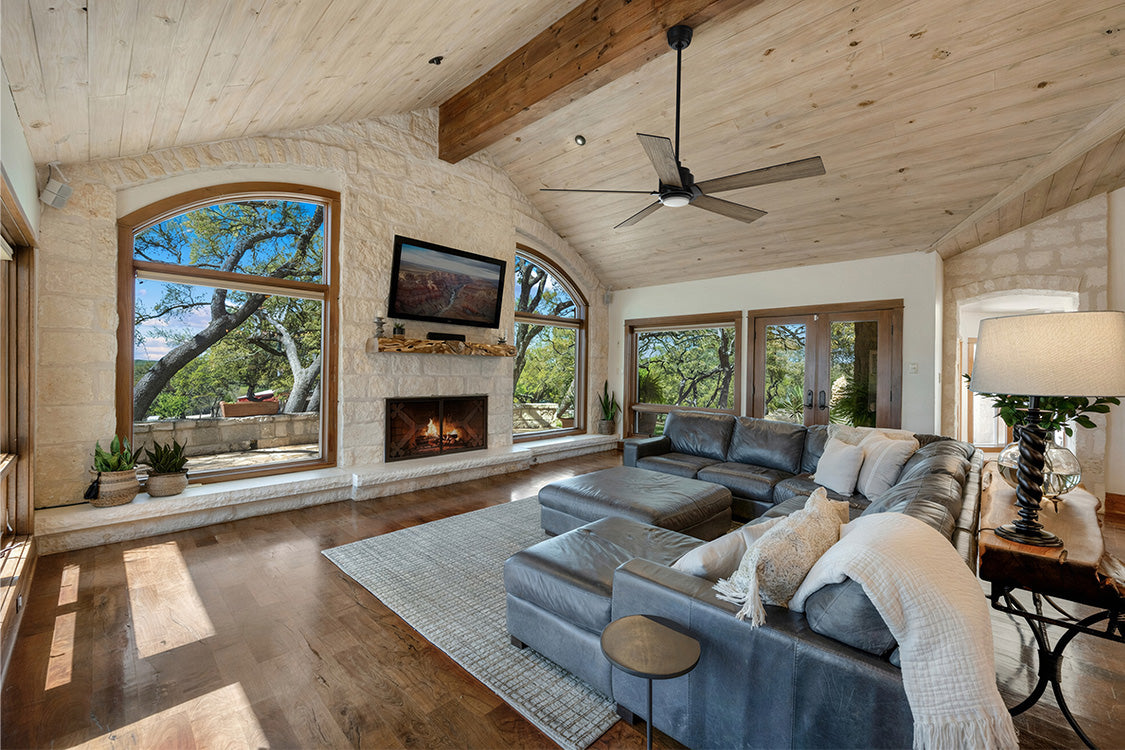 A spacious farmhouse-style living room featuring a vaulted wooden ceiling with exposed beams, a stone fireplace, large windows overlooking an outdoor area, a dark leather sectional sofa, and a black Aspen 60-inch smart ceiling fan mounted overhead.