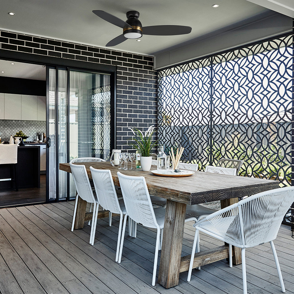A modern outdoor dining area featuring a black Biscay 52-inch outdoor ceiling fan with 3 blades and a modern design, positioned above a rustic wooden dining table with white chairs. The area is enclosed by a decorative privacy screen and has a view into an indoor kitchen.