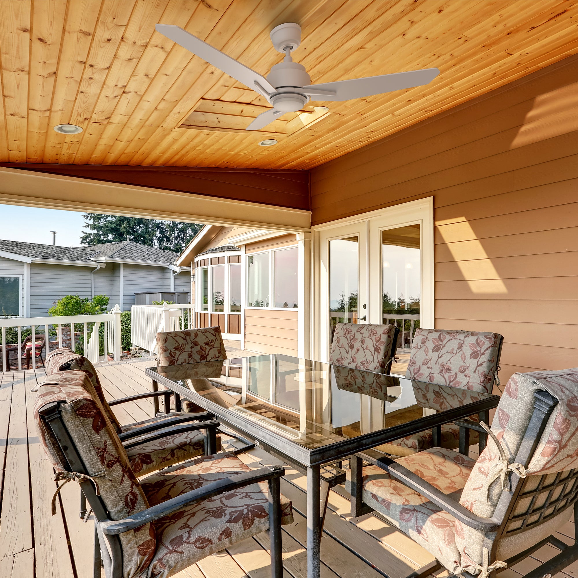 A modern covered patio featuring a 48-inch Carro Smafan Tilbury outdoor smart ceiling fan with LED light and remote, mounted on a white ceiling. The fan, with three sleek blades in a white finish, illuminates a cozy seating area with cushioned wicker chairs, a glass-top coffee table. The patio overlooks a scenic backyard with greenery and wooden fencing, showcasing the fan&
