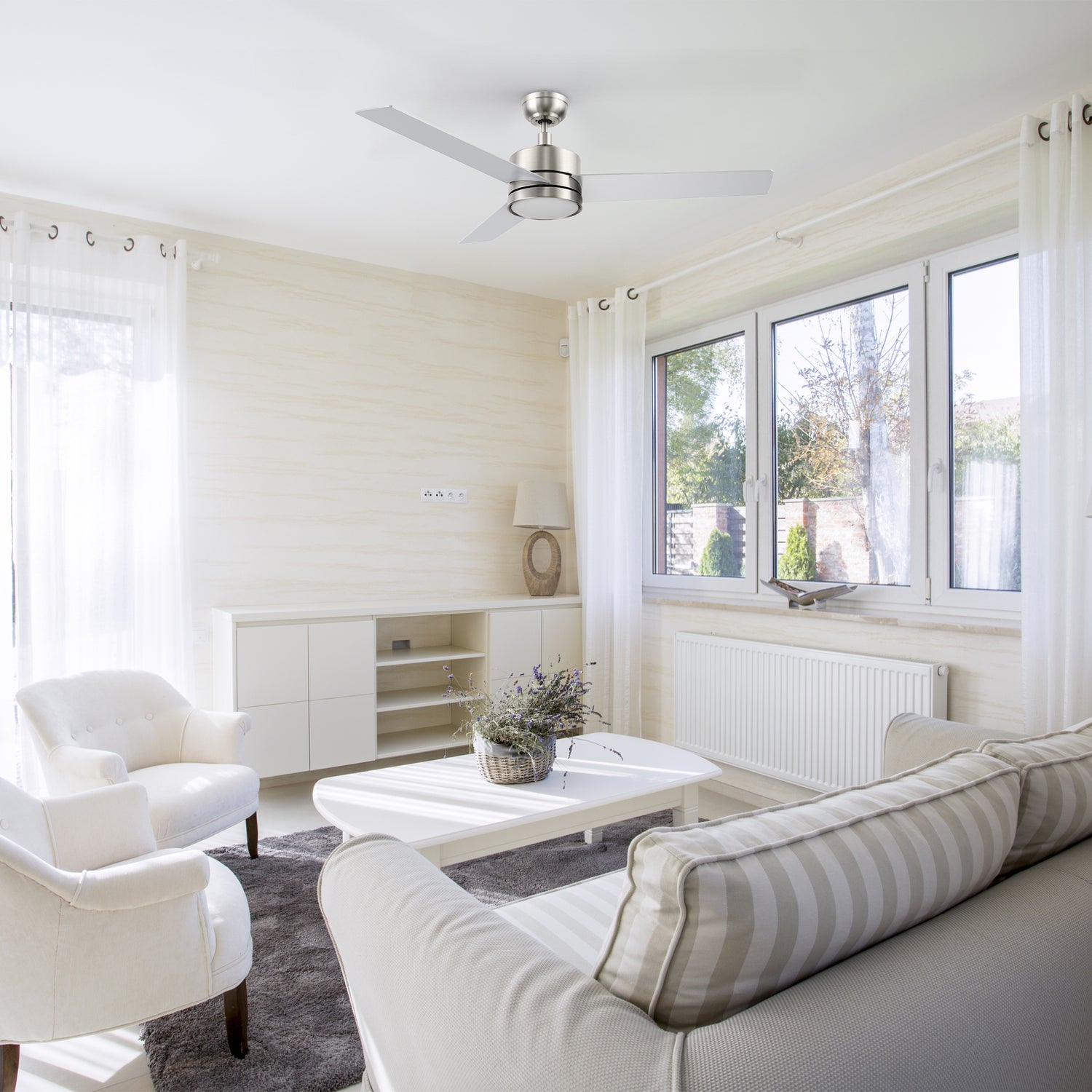 Modern living room featuring the Carro Smafan 52-inch Louy ceiling fan in brushed nickel with integrated LED light, styled in a bright, neutral-toned living room with large windows, white furniture, and soft natural lighting.