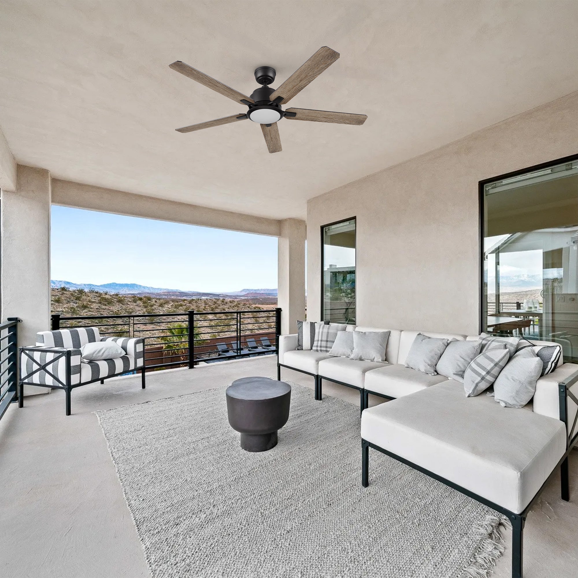 Carro Smafan Essex2 52-inch black smart outdoor ceiling fan with LED light installed on a covered balcony, above a modern outdoor sectional sofa and coffee table, overlooking a scenic view.