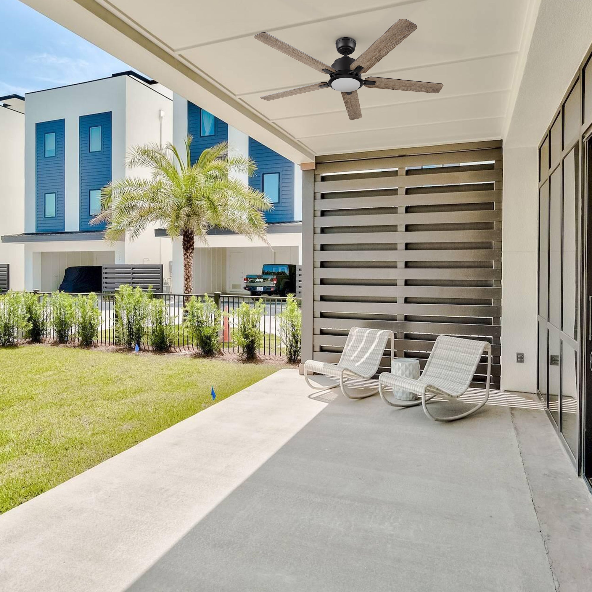 Carro Smafan Essex2 52-inch smart outdoor ceiling fan in black with LED light installed under a covered patio with lounge chairs and tropical landscaping, showcasing an airy and shaded outdoor retreat.