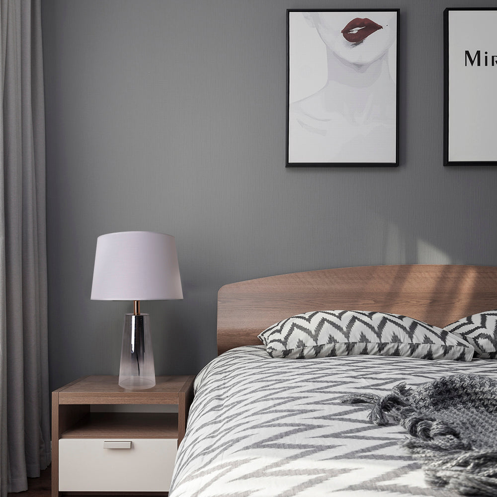 A bedroom side table view of a Carro Smafan Rosemary Table Lamp, 25 inches tall, with a Chrome Grey Ombre finish and white shade, placed on a nightstand. The lamp provides warm lighting, enhancing the room&