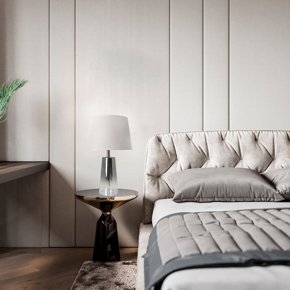 A bedroom view featuring a Carro Smafan Rosemary Table Lamp, 25 inches tall, with a Chrome Grey Ombre finish and white shade, placed on a nightstand beside a bed. The lamp emits a warm glow, complementing the room&