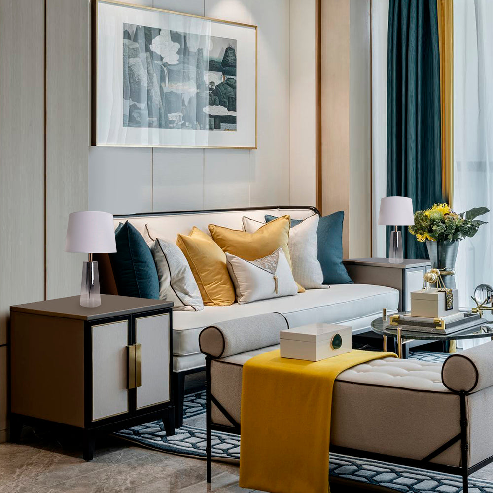 A living room side table view of a Carro Smafan Rosemary Table Lamp, 25 inches tall, with a Chrome Grey Ombre finish and white shade, placed on a table near a sofa. The lamp&