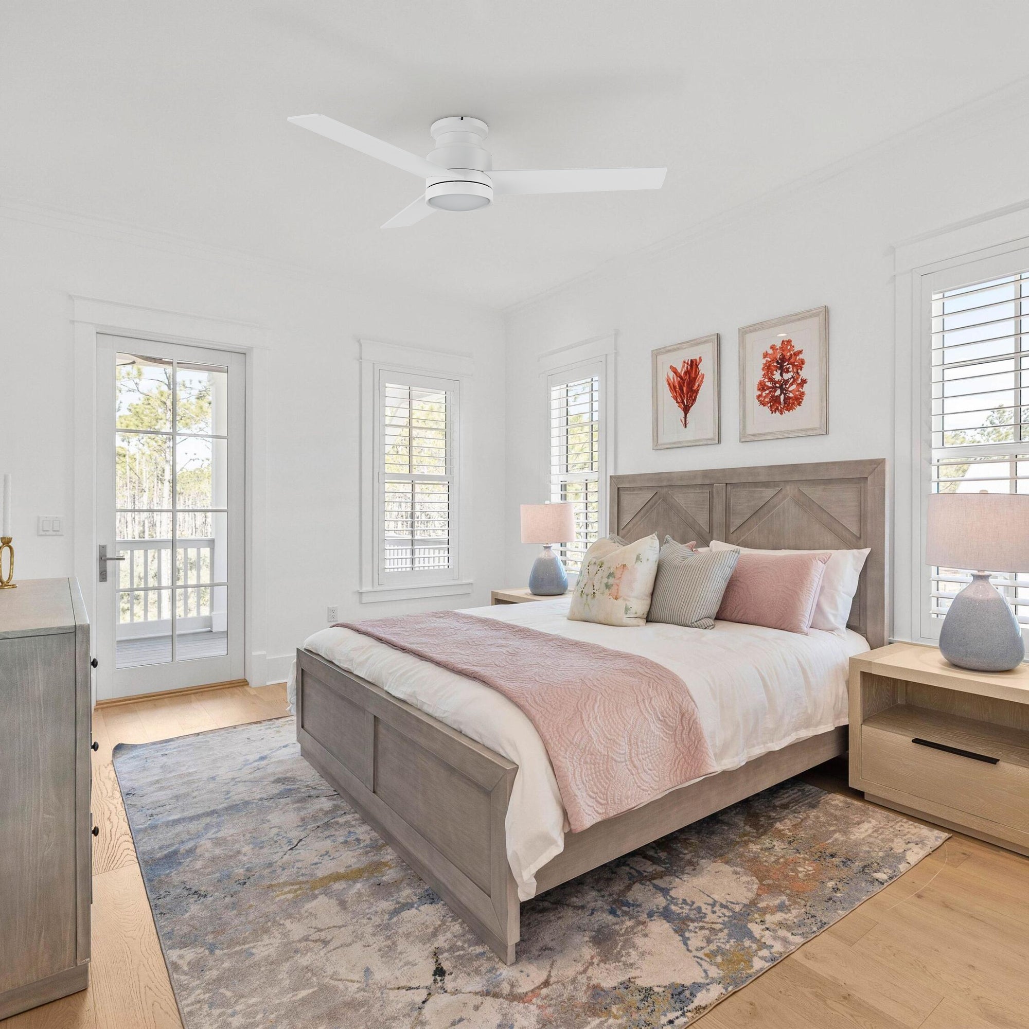 Mid-century bedroom with the Ariccia 52-inch flush mount ceiling fan in pure white, featuring five blades and an integrated LED light. Room includes a wooden bed with pink accents, ceramic lamps on nightstands, a colorful rug, coral wall art, and natural light from shuttered windows.