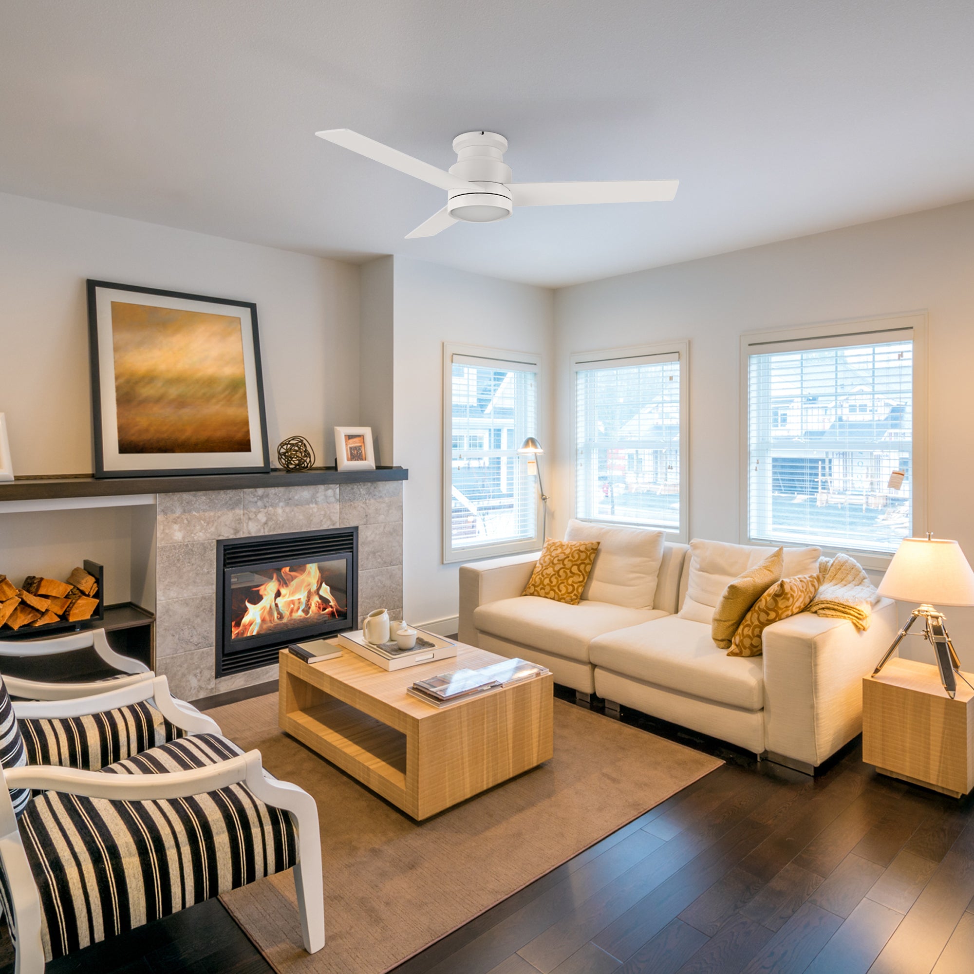 Traditional living room with the Ariccia 52-inch flush mount ceiling fan in pure white, featuring three blades and an integrated LED light. The space includes a stone fireplace, white sofa with yellow cushions, striped armchairs, and large shuttered windows for a warm, inviting atmosphere.