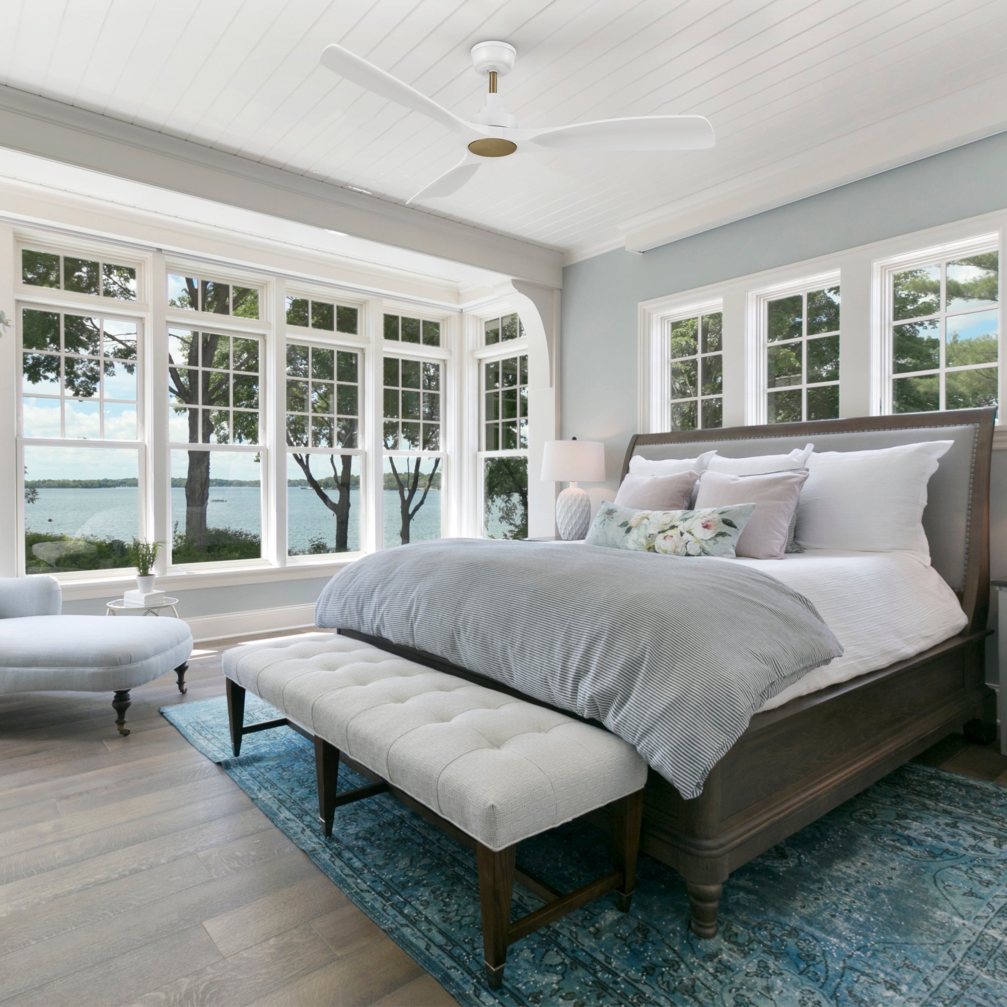 A bedroom with a white smart ceiling fan with gold accents on a white paneled ceiling, enhancing airflow and cooling. Light blue walls, large windows with a lake view, a dark wooden bed with gray bedding, a white tufted bench, and a light blue chair on a light wood floor with a blue rug.