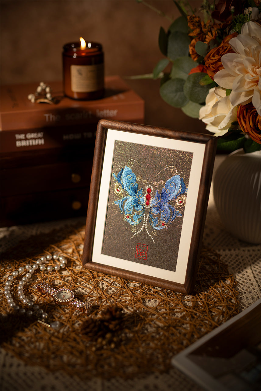 A wood-framed embroidered artwork showing beautiful blue butterfly, paired with flowers and jewelry and scented candles, is placed on the tabletop.