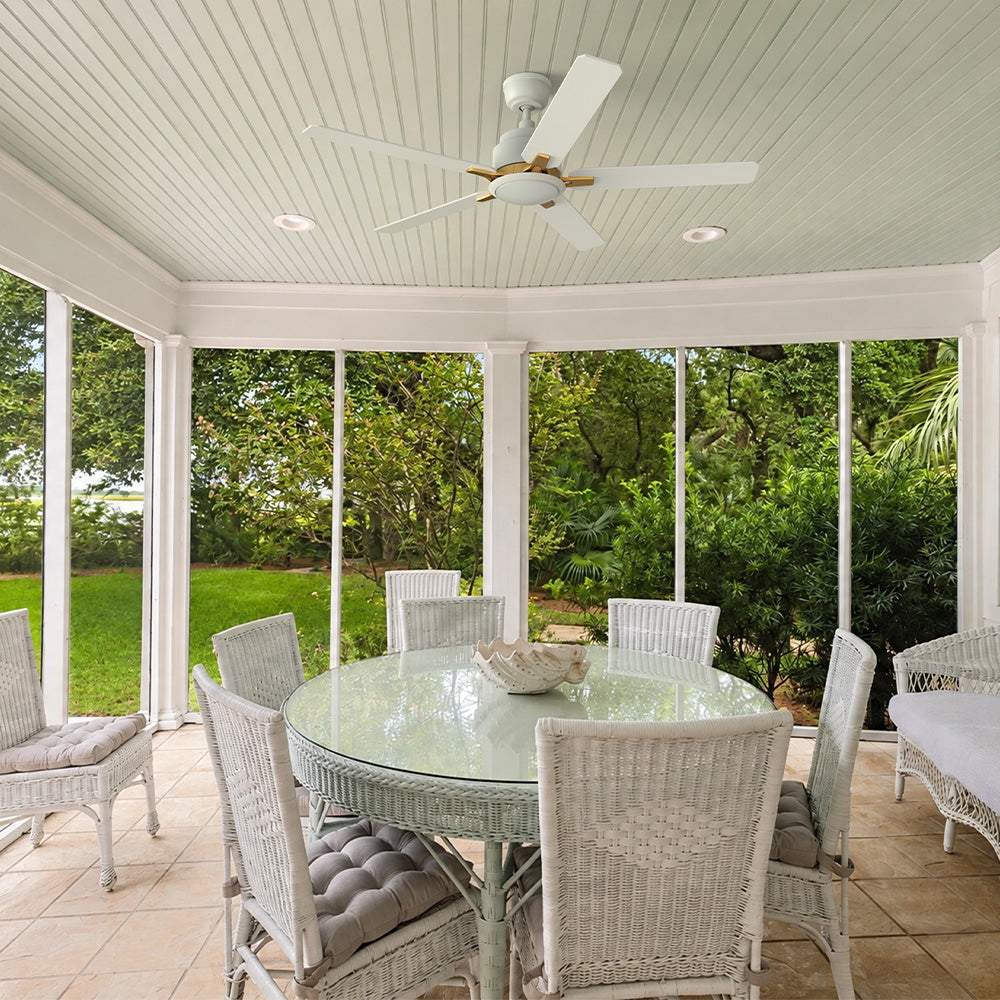 A covered patio with a white ceiling features the Essex 52" White Outdoor Ceiling Fan with gold-accented blades. Paired with a wicker dining set and lush greenery, it brings cool comfort and elegant style to outdoor living.