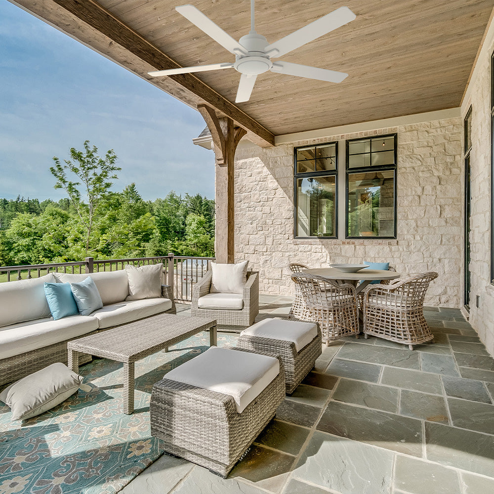 A white, five-blade outdoor ceiling fan is mounted to the wooden ceiling of a spacious, covered stone patio. The patio is furnished with a modern wicker sectional, matching armchairs, and a small dining set, all overlooking a lush green forest and a body of water under a bright blue sky.