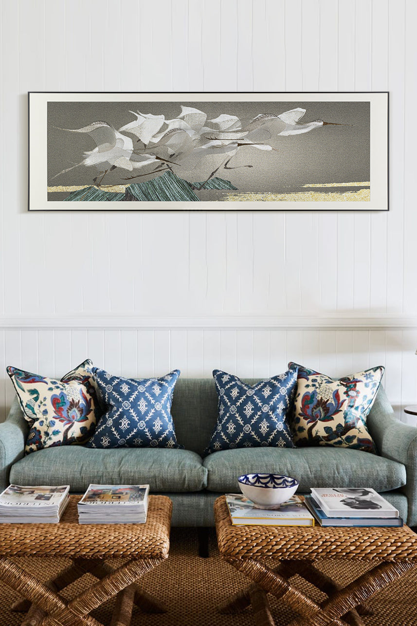 A framed embroidery wall art piece depicting egrets flying over mountains, displayed above an elegant sofa with blue and white patterned cushions in front of it. The room features wicker coffee tables adorned with magazines and vases, all set against pristine white panelled walls.