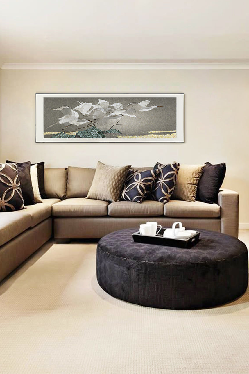 A framed embroidery artwork of egrets hanging in a family room with beige sofas and a round ottoman.