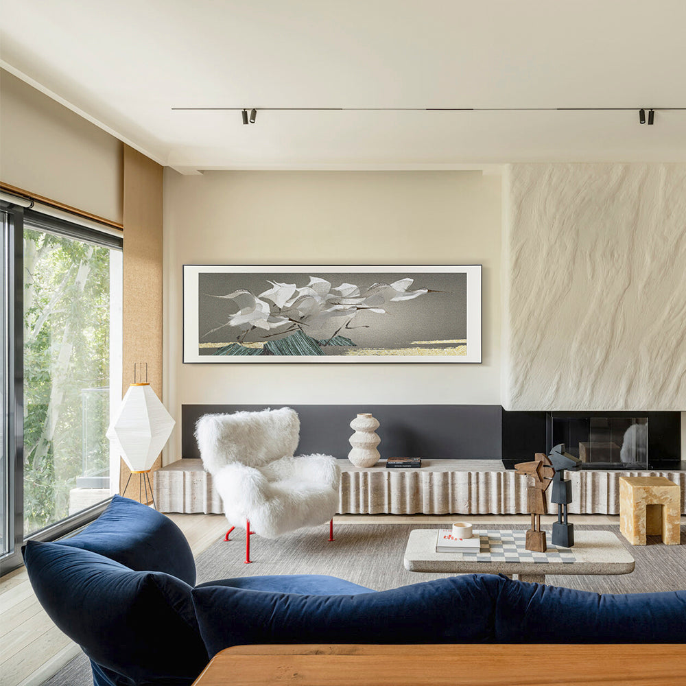 A framed embroidery artwork of egrets displayed in a modern living room with a large window and blue furniture.