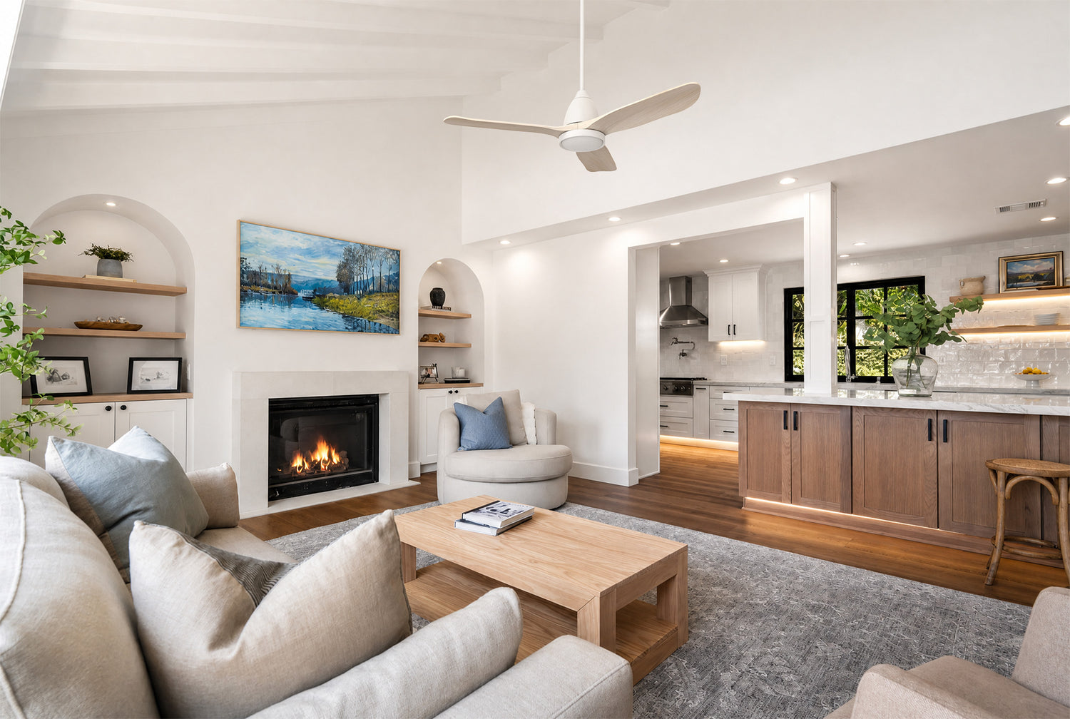 Smafan white ceiling fan with light 3 wood blades installed on a vaulted ceiling in a bright open-concept modern living room with fireplace and kitchen.