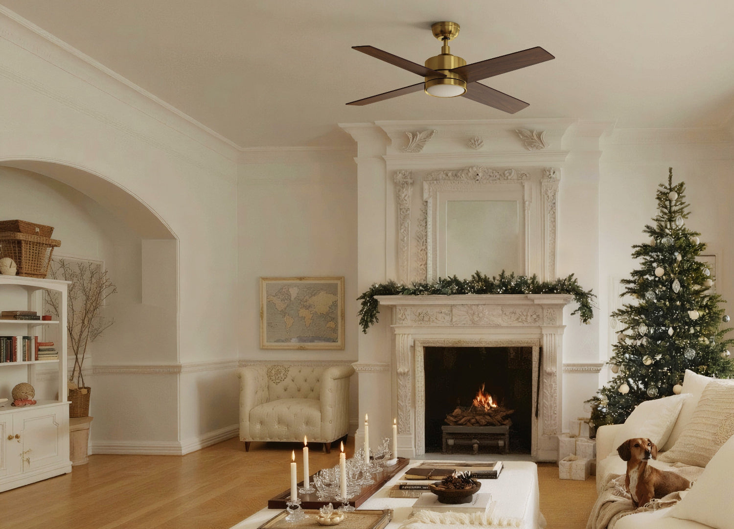 A festive living room decorated for Christmas with a lit tree, a dog on a white couch, a fireplace with a garland and candles, and Golg high-end ceiling fan with light overhead.