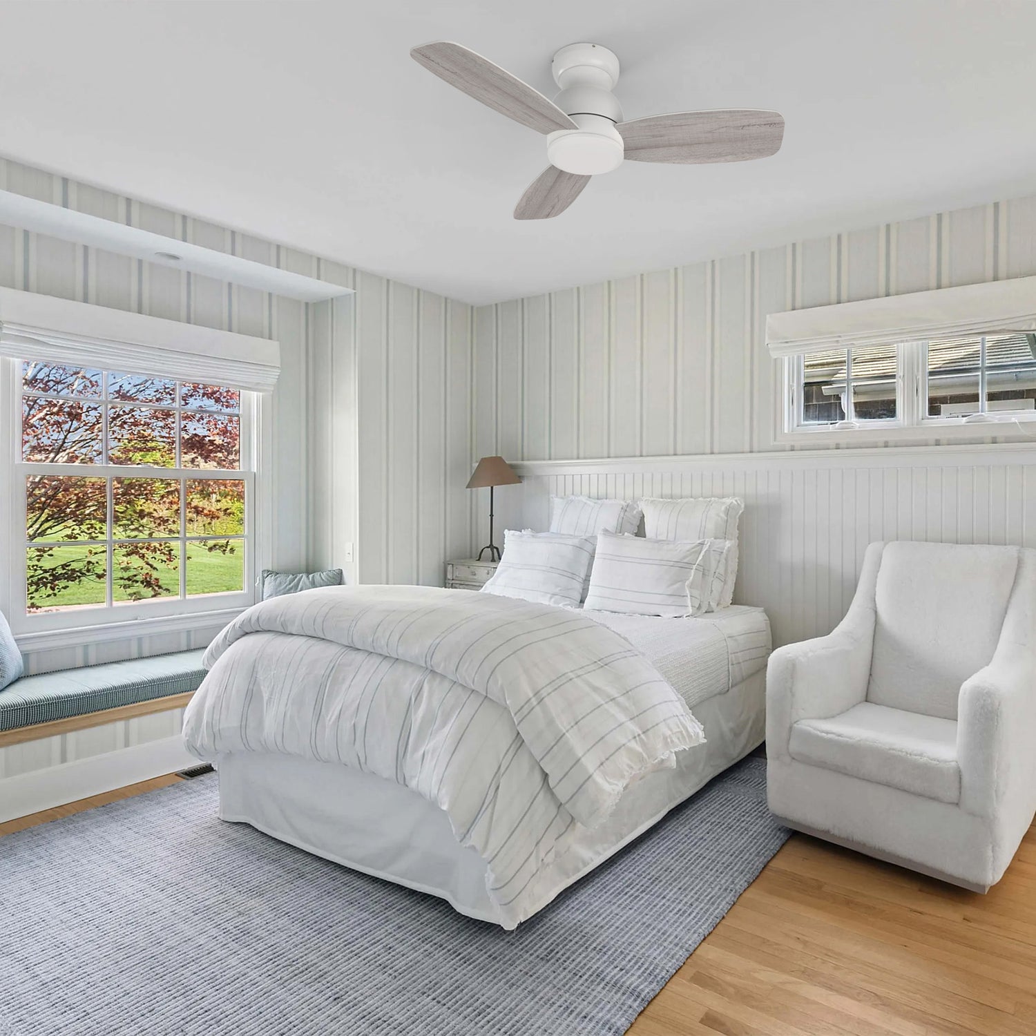 A serene bedroom featuring a CARRO USA Osborn 44-in flush mount white ceiling fan without a light kit, positioned above a bed with white and blue striped bedding. The room includes a large window with a built-in window seat, a white armchair, and light-colored wooden floors with a blue patterned rug.