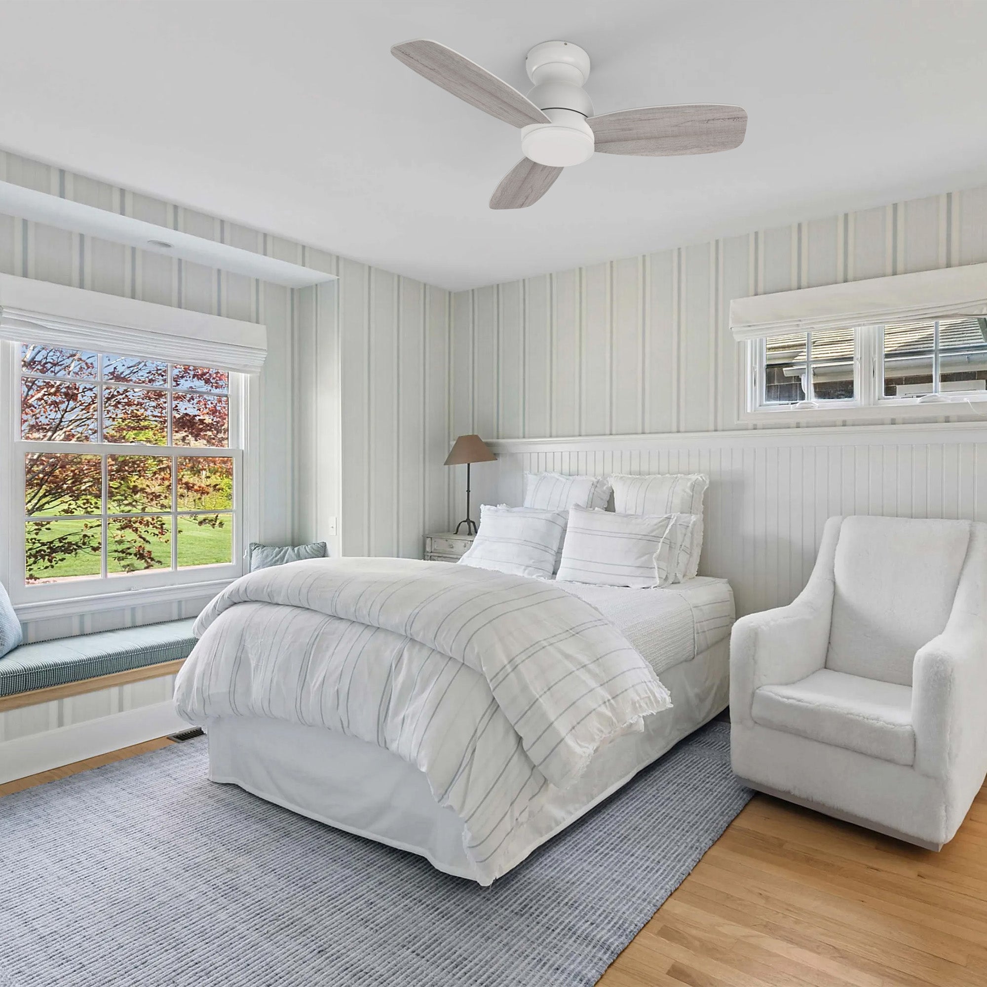 A serene bedroom featuring a CARRO USA Osborn 44-in flush mount white ceiling fan without a light kit, positioned above a bed with white and blue striped bedding. The room includes a large window with a built-in window seat, a white armchair, and light-colored wooden floors with a blue patterned rug.