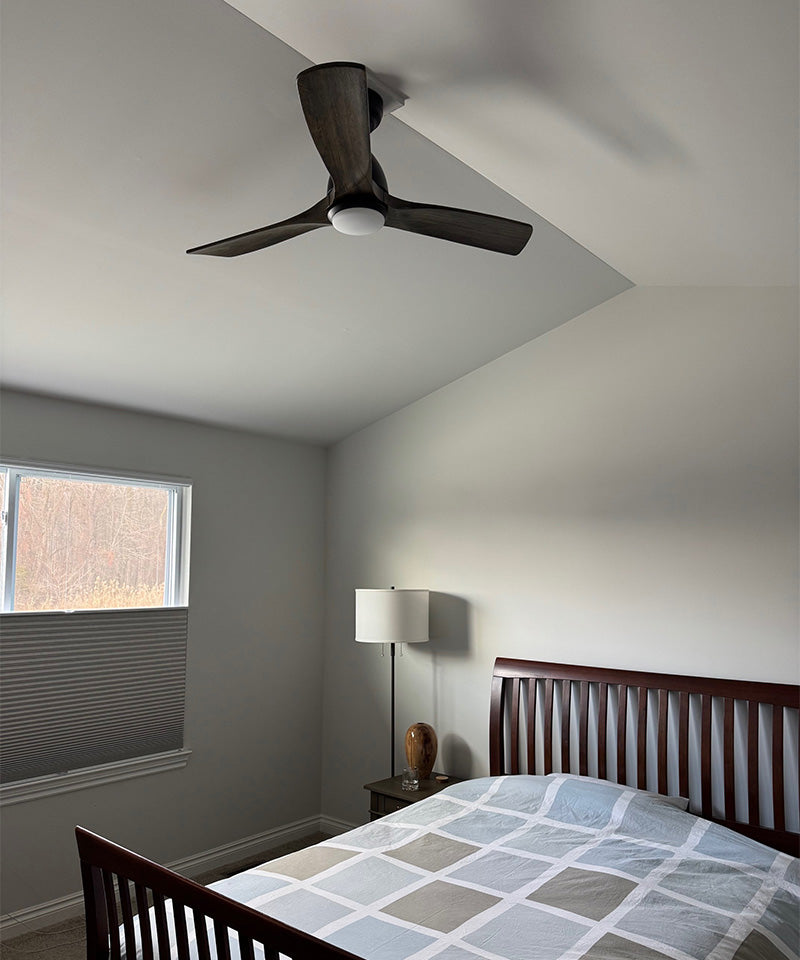 A cozy bedroom with light gray walls and a vaulted ceiling featuring a Smafan Sawyer 48-inch smart wood ceiling fan with light. The dark wood, three-blade fan hangs elegantly above a matching dark wood bed frame dressed in striped bedding. A nightstand with a lamp sits to the right of the bed, while a window with a cellular shade allows soft natural light into the room.