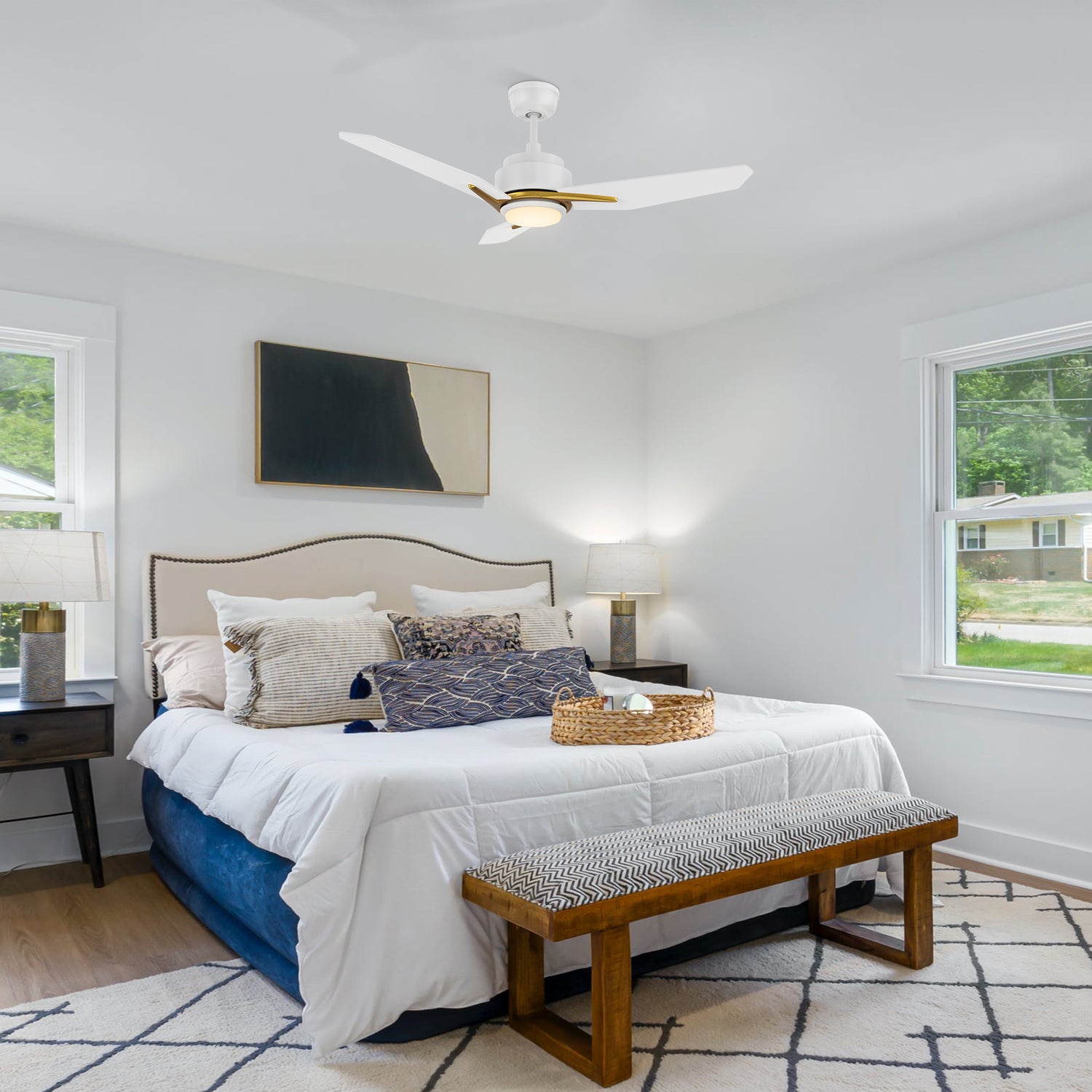 The image shows a modern bedroom with a white and gold ceiling fan with a light.  The bed has a beige headboard, decorated with various neutral and blue pillows, and a white comforter, with a woven basket containing a plant.  Matching nightstands with lamps are on either side, and a dark wood bench with a patterned cushion sits at the foot.  The room features light-colored walls, two windows with natural light, an abstract painting above the bed, and a patterned rug on the wooden floor.