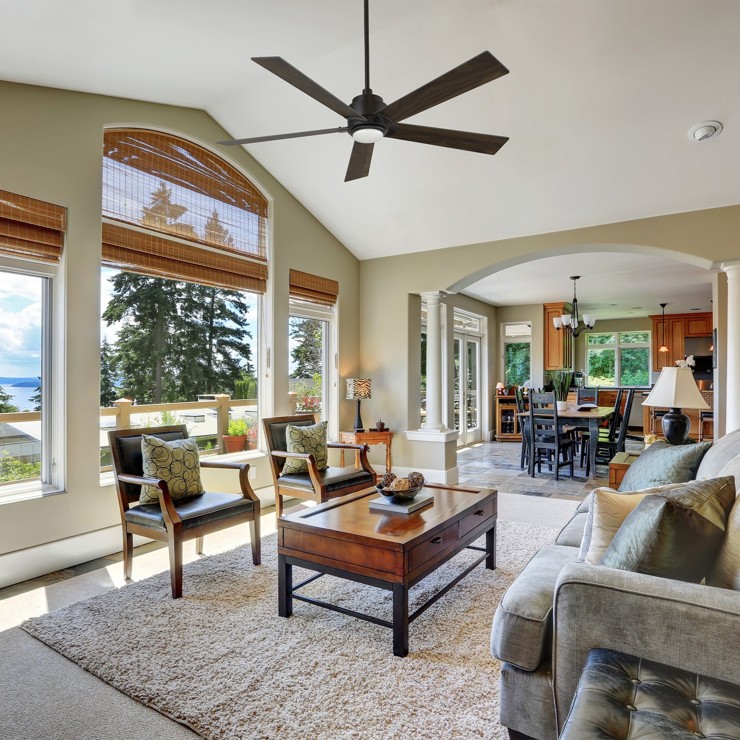 Kalmar 60-inch remote control ceiling fan with LED light installed in a bright living room featuring large windows, wooden furniture, and an open-concept layout connecting to the dining area.