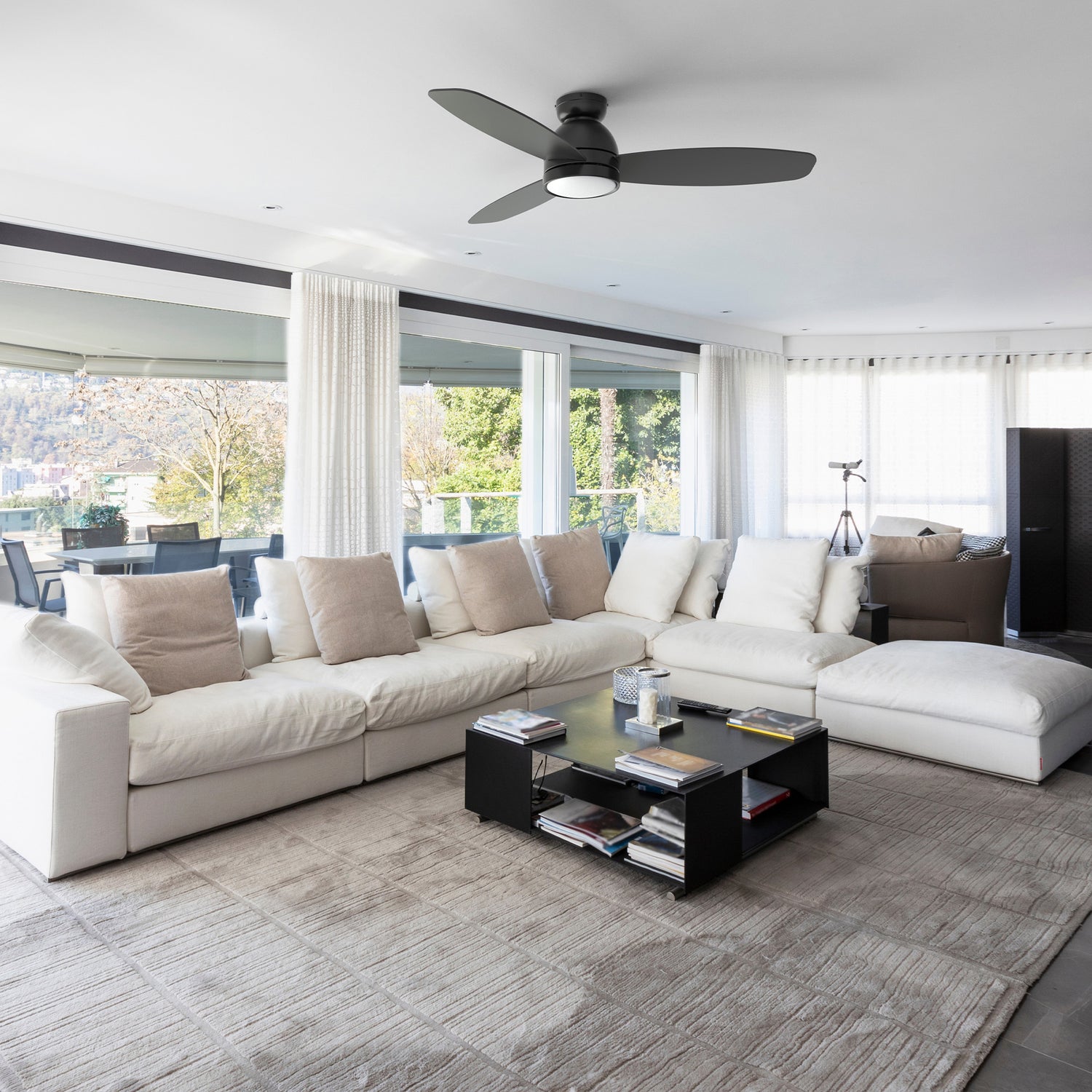 A modern living room features a large, off-white sectional sofa and a dark rectangular coffee table on a textured grey rug. In the center of the ceiling, a black, modern, 52 inch flush-mount ceiling fan with integrated light is visible. Large windows overlook an outdoor area with trees and a table and chairs.