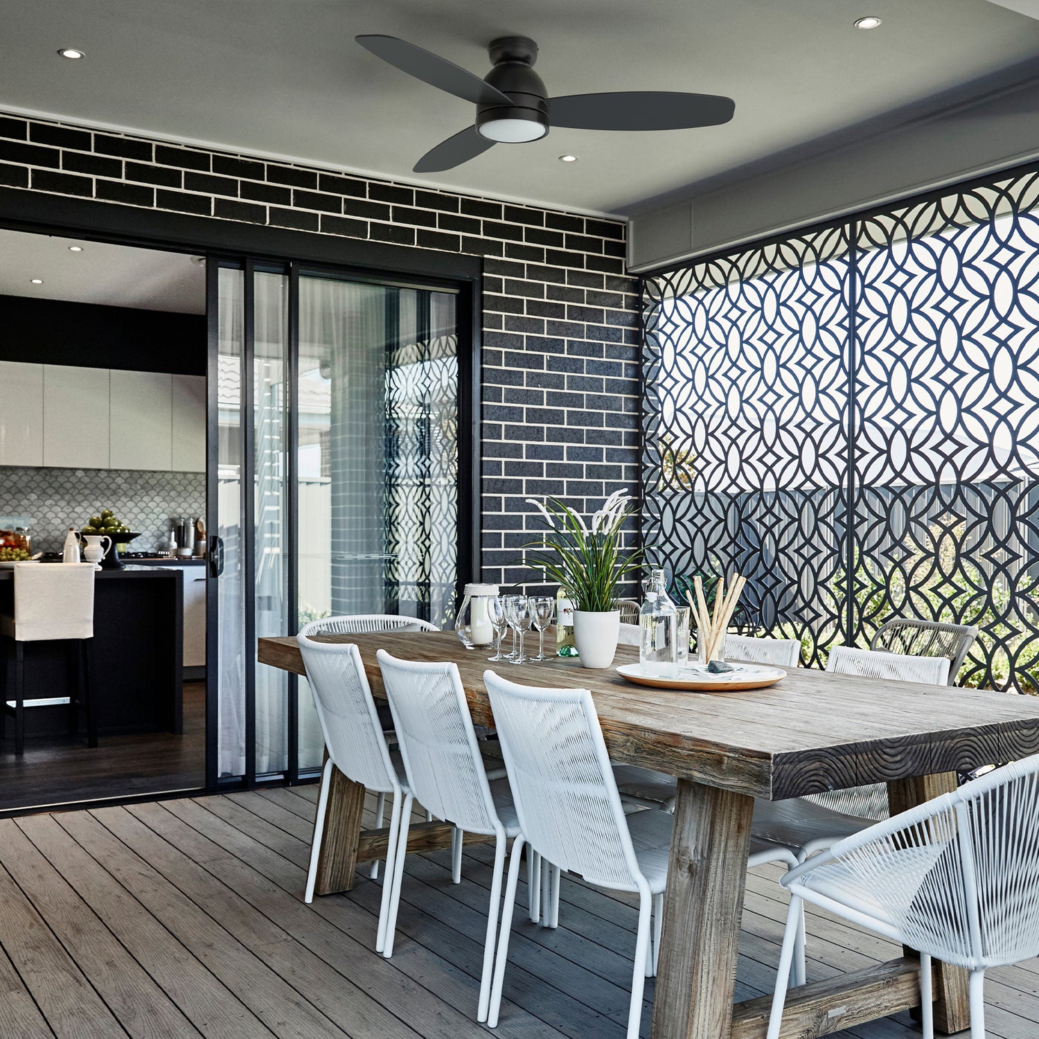 Modern outdoor dining area with Smafan Biscay 52-inch low profile black ceiling fan with light and smart, mounted above a rustic wooden table and white chairs, featuring decorative privacy screens and sliding glass doors leading to the kitchen.
