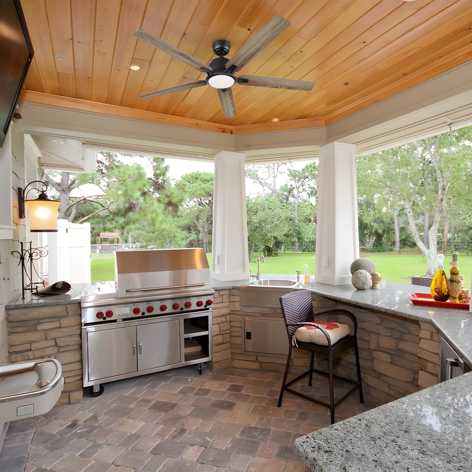 An outdoor kitchen with natural stone and granite finishes, featuring the 56-inch Smafan Essex smart ceiling fan in a walnut finish. The fan is mounted on a wood-paneled ceiling and includes an integrated LED light. The space is equipped with a built-in stainless steel grill, bar-style seating, and large open windows overlooking a lush green lawn, making it ideal for outdoor entertaining.