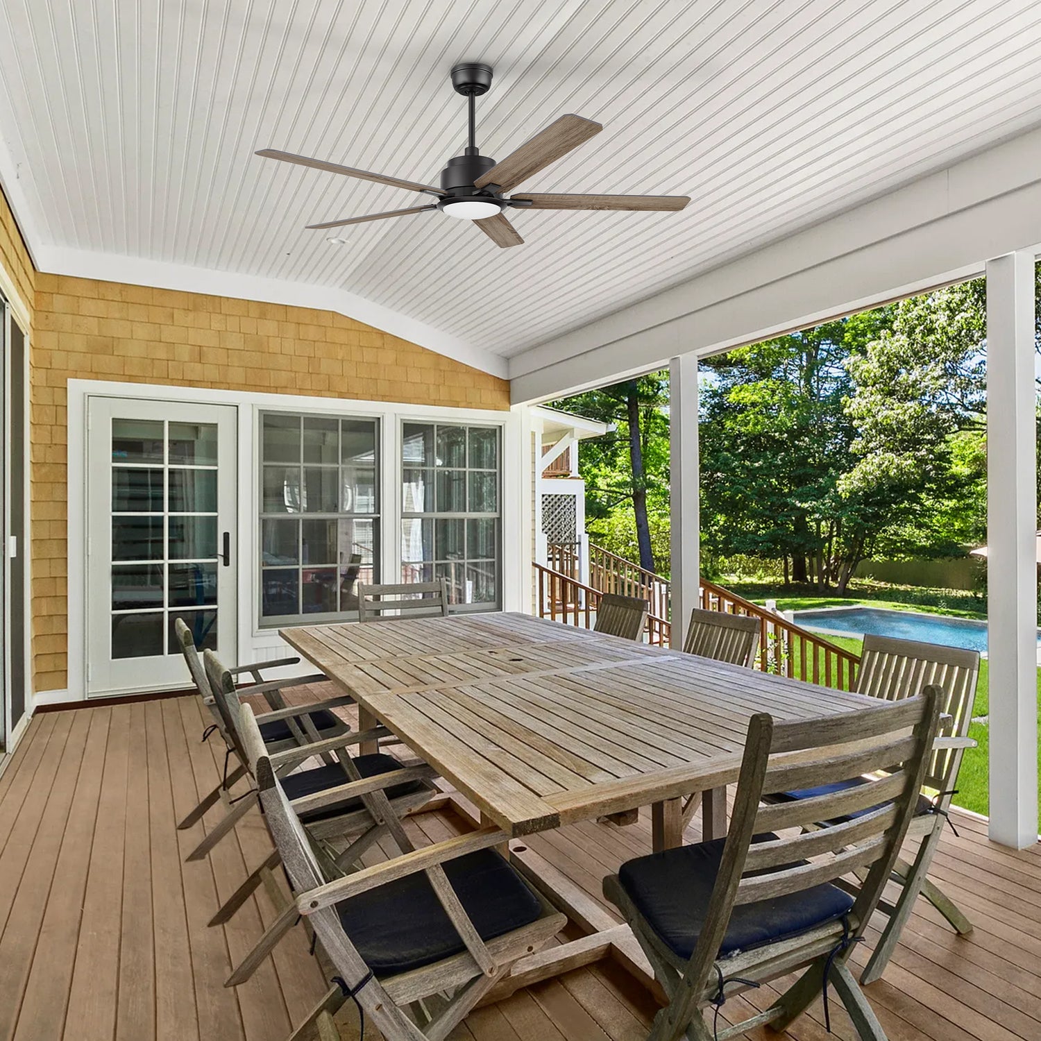 A spacious covered patio with a long wooden dining table and eight chairs, featuring the 56-inch Smafan Essex outdoor smart ceiling fan in walnut finish. The fan is mounted on a white paneled ceiling, providing cooling and lighting for the outdoor dining area. The space overlooks a backyard with lush greenery and a swimming pool.