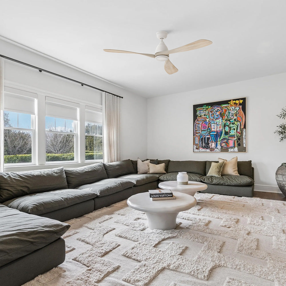 White 60 inch 3-blade light wood ceiling fan with LED light installed on the ceiling of an organic modern living room with a large sectional sofa.