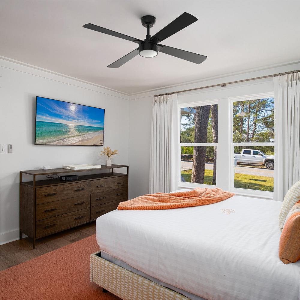 A Smafan Admiral 52-inch matte black 4-blade ceiling fan with an integrated LED light kit, installed on a white bedroom ceiling. The modern bedroom features a bed with white bedding and an orange throw blanket, a peach-colored area rug, a dark wood dresser, a wall-mounted TV showing a beach scene, and large windows with white curtains looking outside.