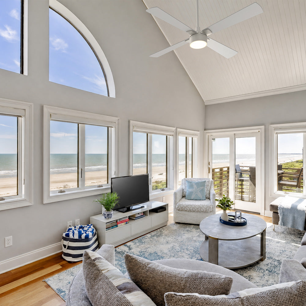 White Smafan Admiral 52-inch ceiling fan with light installed on a high vaulted ceiling in a coastal living room with large ocean-view windows.