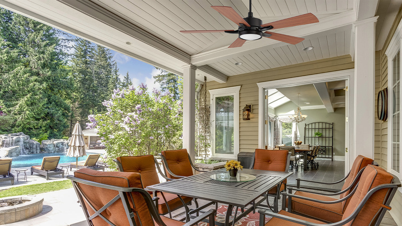 Outdoor patio scene with a 52-inch black smart ceiling fan with light and wooden blades hanging from the top. The patio features brown cushioned chairs and a black table with a yellow flower vase. The background includes green trees, purple flowering bushes, a swimming pool with a waterfall, and a white house under a blue sky. The floor is gray tiled, with a fire pit on the left and an indoor view on the right.