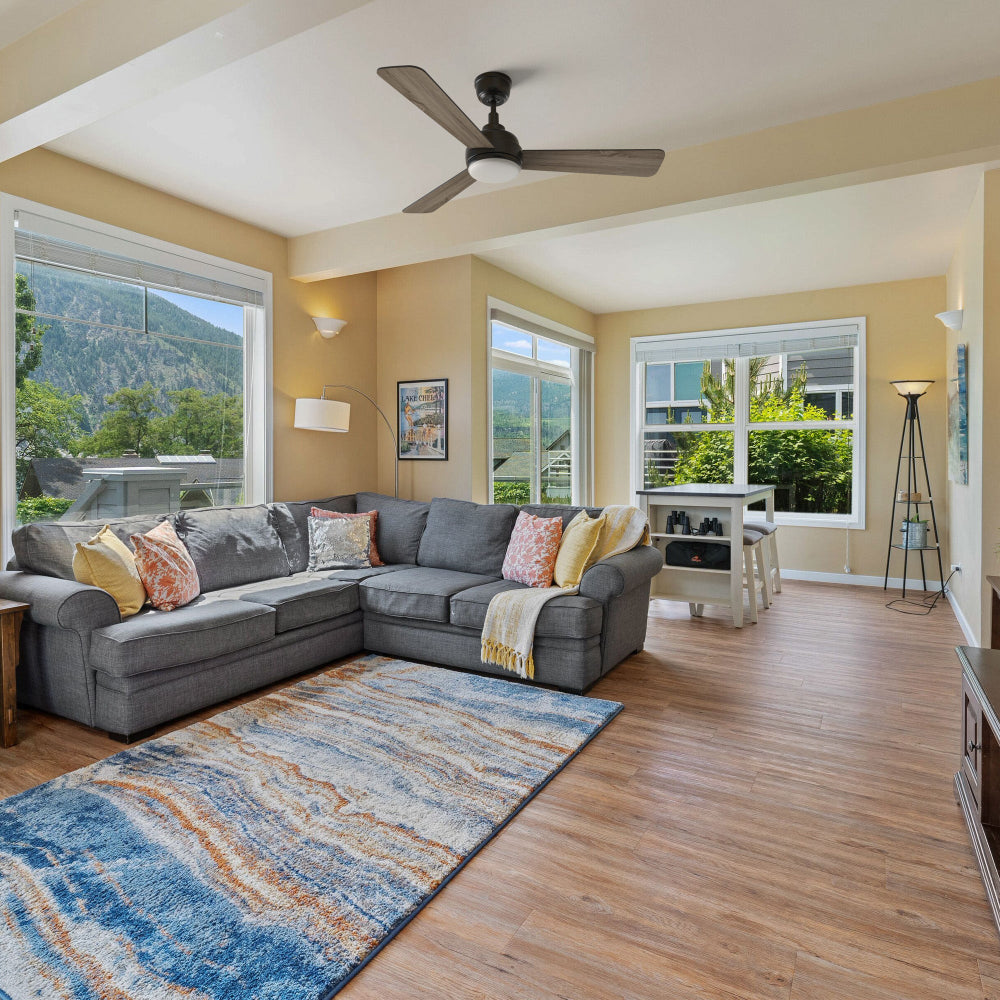 A bright modern Bohemian living room with a gray sectional sofa, colorful pillows, and a yellow throw, centered around a blue and orange area rug on warm wood flooring. Overhead, the Smafan Audra 52-inch 3-blade ceiling fan with light provides airflow and illumination. Large windows frame mountain views, enhancing the inviting space.