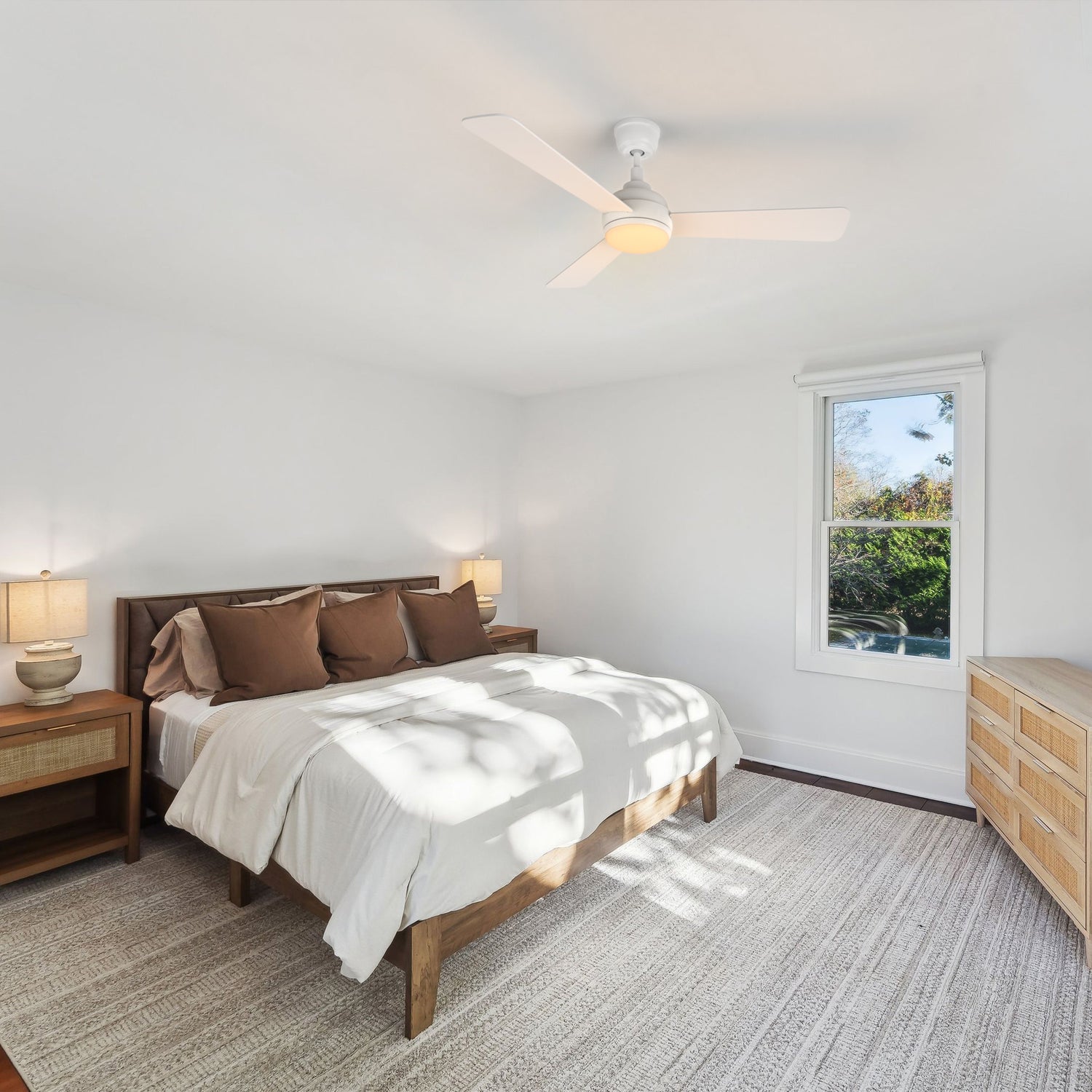 An inviting rustic bedroom with a neatly made bed, brown headboard, white duvet, and brown pillows rests on a light gray rug. Overhead, a white Smafan Audra 52-inch ceiling fan with 3 blades and an integrated dimmable light provides airflow and illumination. A wooden nightstand with a lamp sits on the left, while sunlight highlights a dresser on the right.