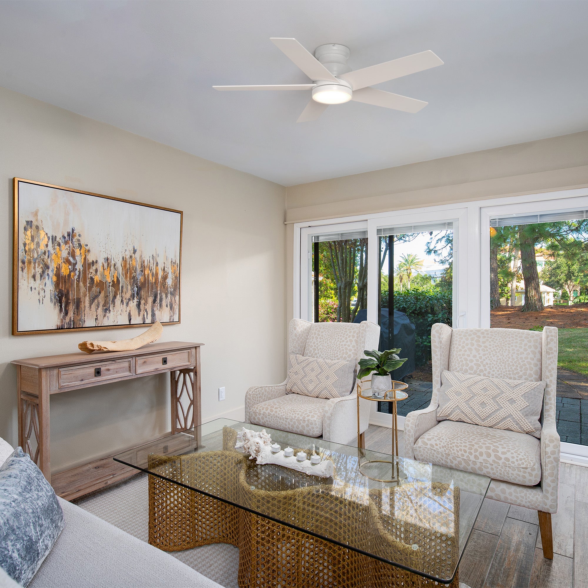 Smafan Bayu 52-inch white low-profile ceiling fan with light in a bright coastal-traditional living room featuring sofa, patterned armchairs, glass-top coffee table, wooden console, and large windows with garden view.