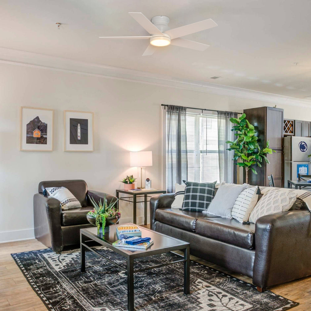 Modern living room with brown leather sofa and armchair, patterned rug, and Smafan Bayu 52-inch white low-profile ceiling fan with light. Dining area visible in background with large plant near window.