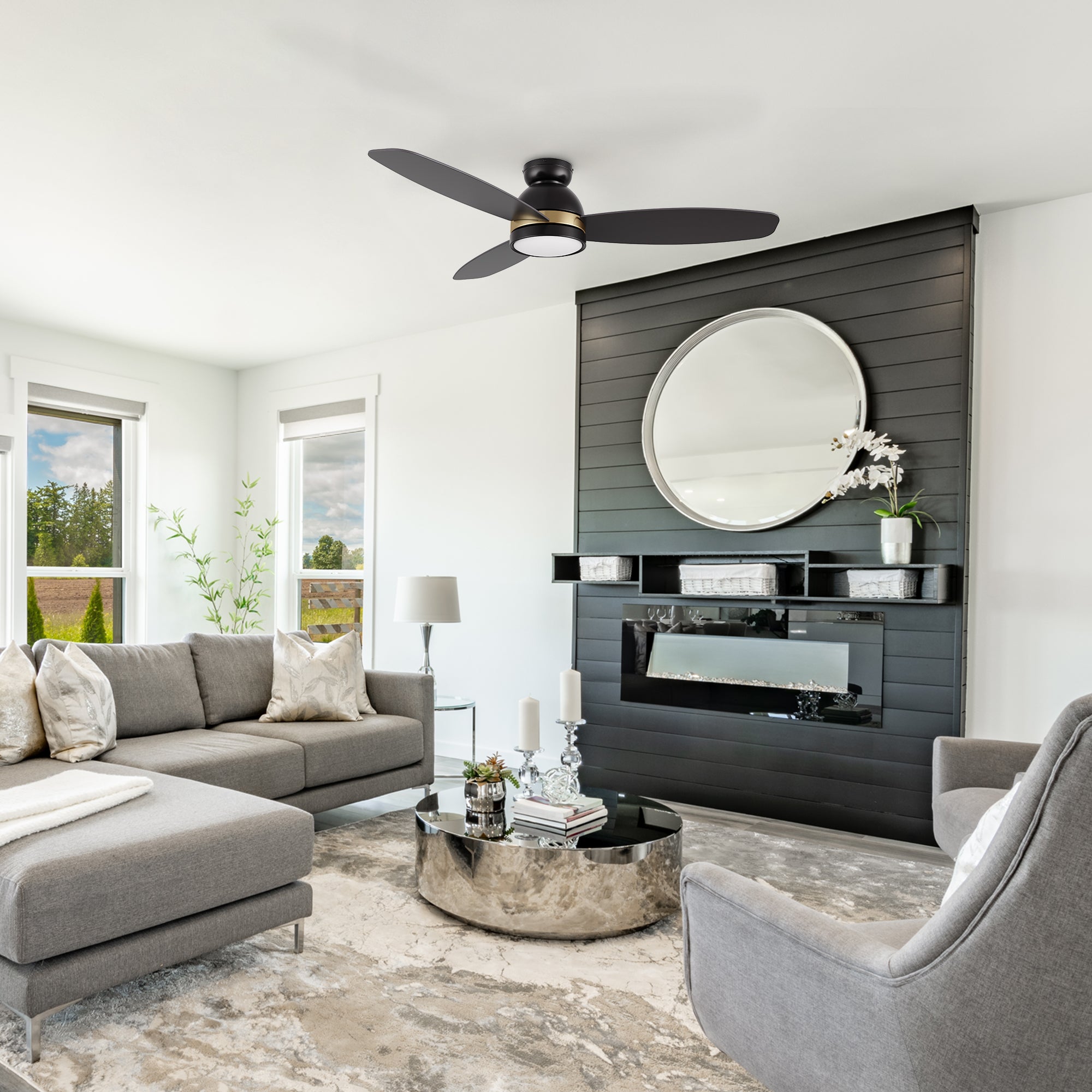 Modern living room featuring the Smafan Biscay 48-inch smart ceiling fan in black with integrated light. The space includes a grey sectional sofa, round mirrored coffee table, and a black accent wall with a circular mirror and electric fireplace.