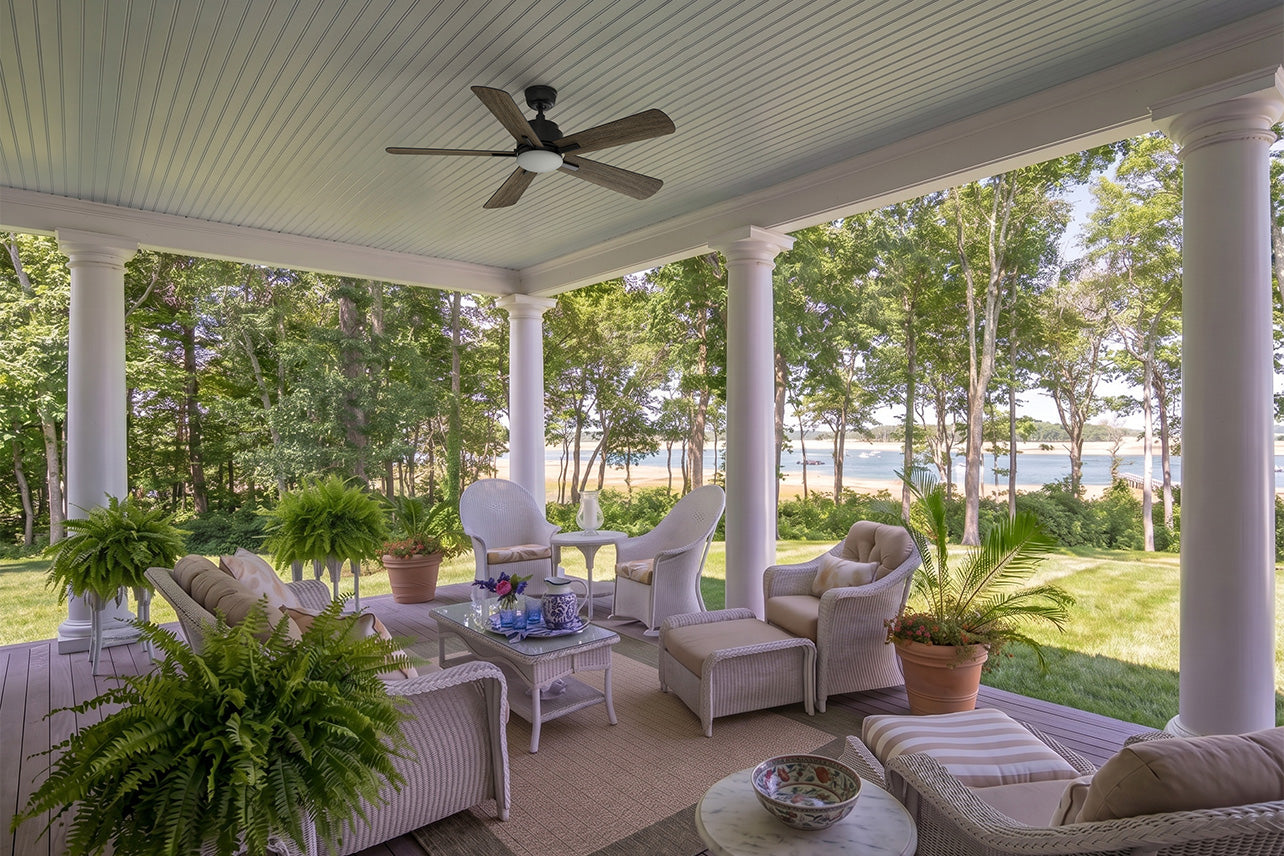 Smafan Elgin outdoor ceiling fan with light installed on a spacious covered porch overlooking a waterfront landscape. The black ceiling fan features five wood-tone blades and an integrated LED light, complementing the traditional coastal-style outdoor living area. White wicker seating, lush greenery, and scenic water views create a relaxing space ideal for patios, porches, and outdoor entertaining.