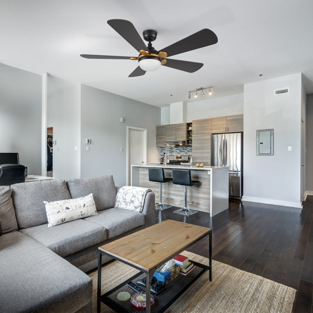 Smafan Lille 52 inch unique ceiling fan in black and gold with light fixture is centered in a modern living room and kitchen area. The living room features a large gray sectional sofa with a patterned pillow and a throw blanket, a wooden coffee table with a metal base, and a woven rug. The kitchen, visible in the background, has a white island with two black bar stools, light wood cabinetry, a stainless steel refrigerator, and tiled backsplash.