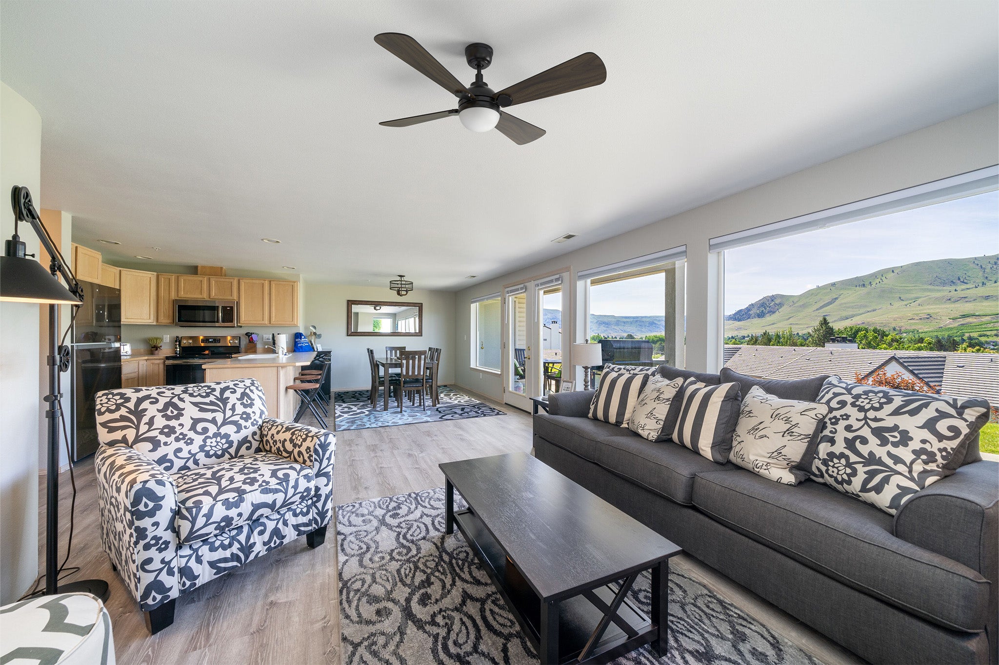 Smafan Rinard 52 inch ceiling fan with LED light, modern farmhouse living room featuring light wood floors, dark gray sofa, patterned armchair, wood dining table, and large windows with scenic valley views.