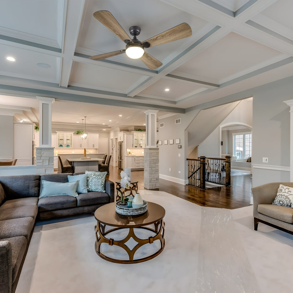 A spacious, open-concept living room with a coffered ceiling and a large ceiling fan with bright light and wooden blades. The room features a dark sectional sofa with light blue throw pillows, a round wooden coffee table, and a light-colored armchair. In the background, an open kitchen with white cabinetry and an island is visible, along with a staircase leading to an upper level.