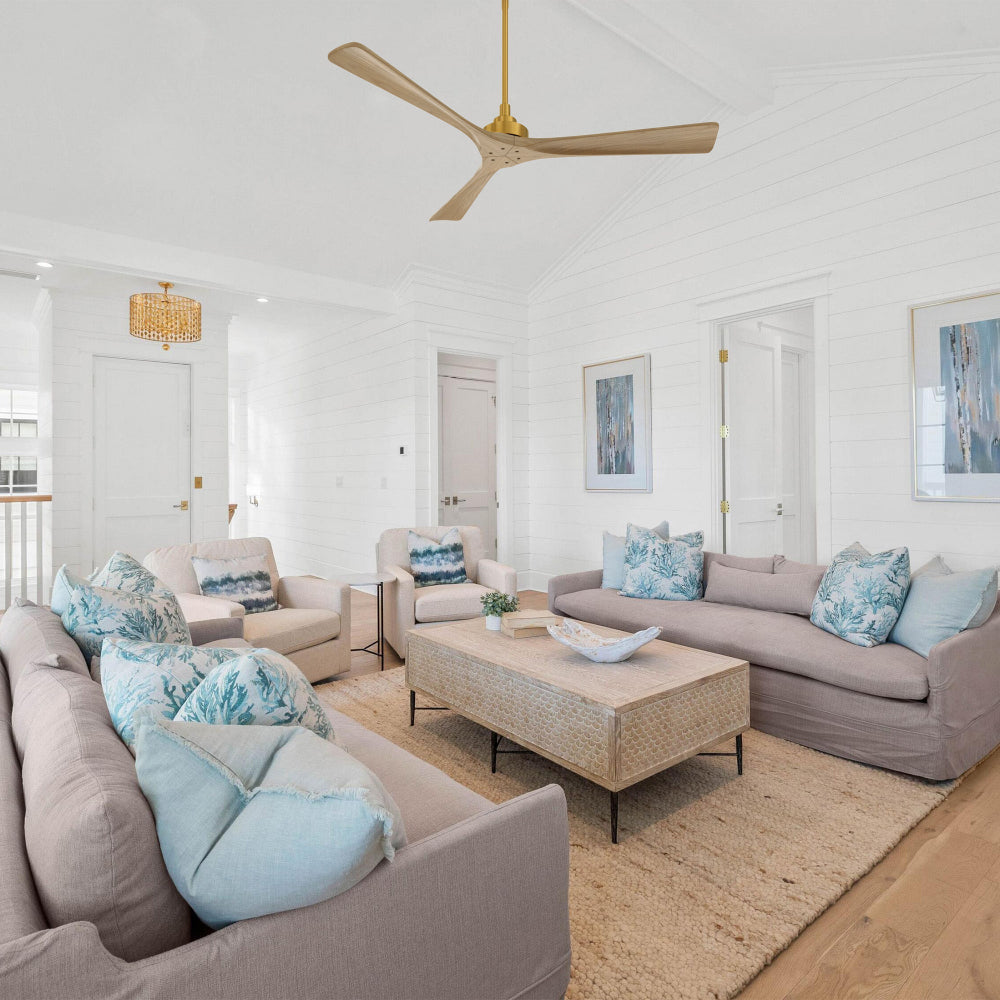 Smafan Thora 60 inch wood ceiling fan with remote in a gold and wood finish, installed in a bright coastal traditional living room with white shiplap walls, light sofas with blue patterned pillows, a woven coffee table on a jute rug, and blue-toned wall art for a relaxed, airy style.