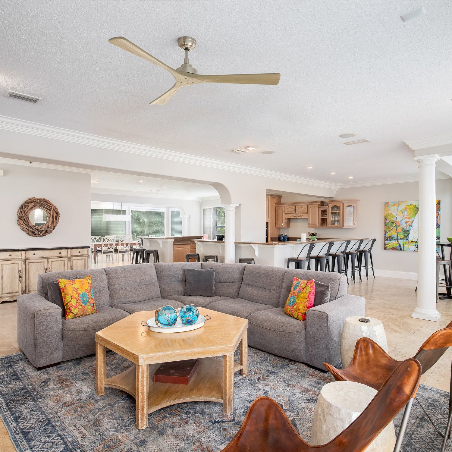 Smafan Thora 60 inch wood ceiling fan with remote in a spacious open-concept transitional living room featuring a gray L-shaped sectional sofa with colorful pillows, light wood octagonal coffee table, brown leather butterfly chairs, and a modern kitchen with breakfast bar in the background.