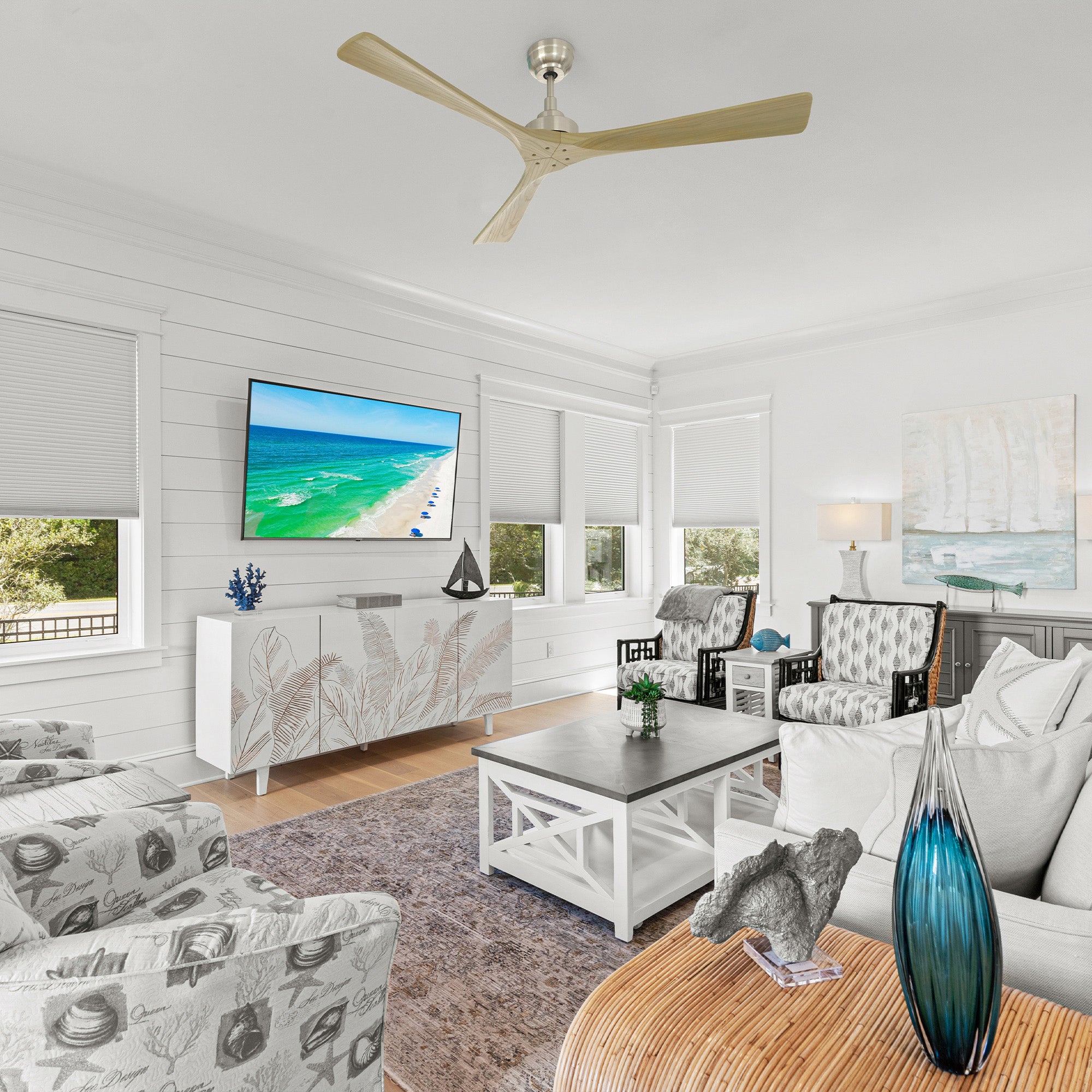 Smafan Thora 60 inch wood ceiling fan with remote in a bright coastal-style living room featuring a large TV above a white leaf-pattern credenza, white and dark gray coffee table, patterned armchairs, and a cozy sofa.